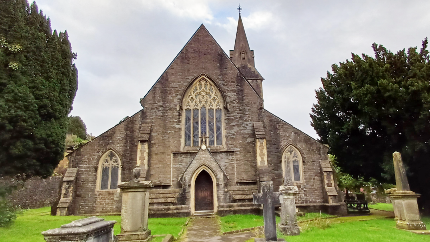 St Mary the Virgin Church has three stained glass windows around the entrance at the front, with a steeple at the back. A number of gravestones can be seen, along with two large trees.