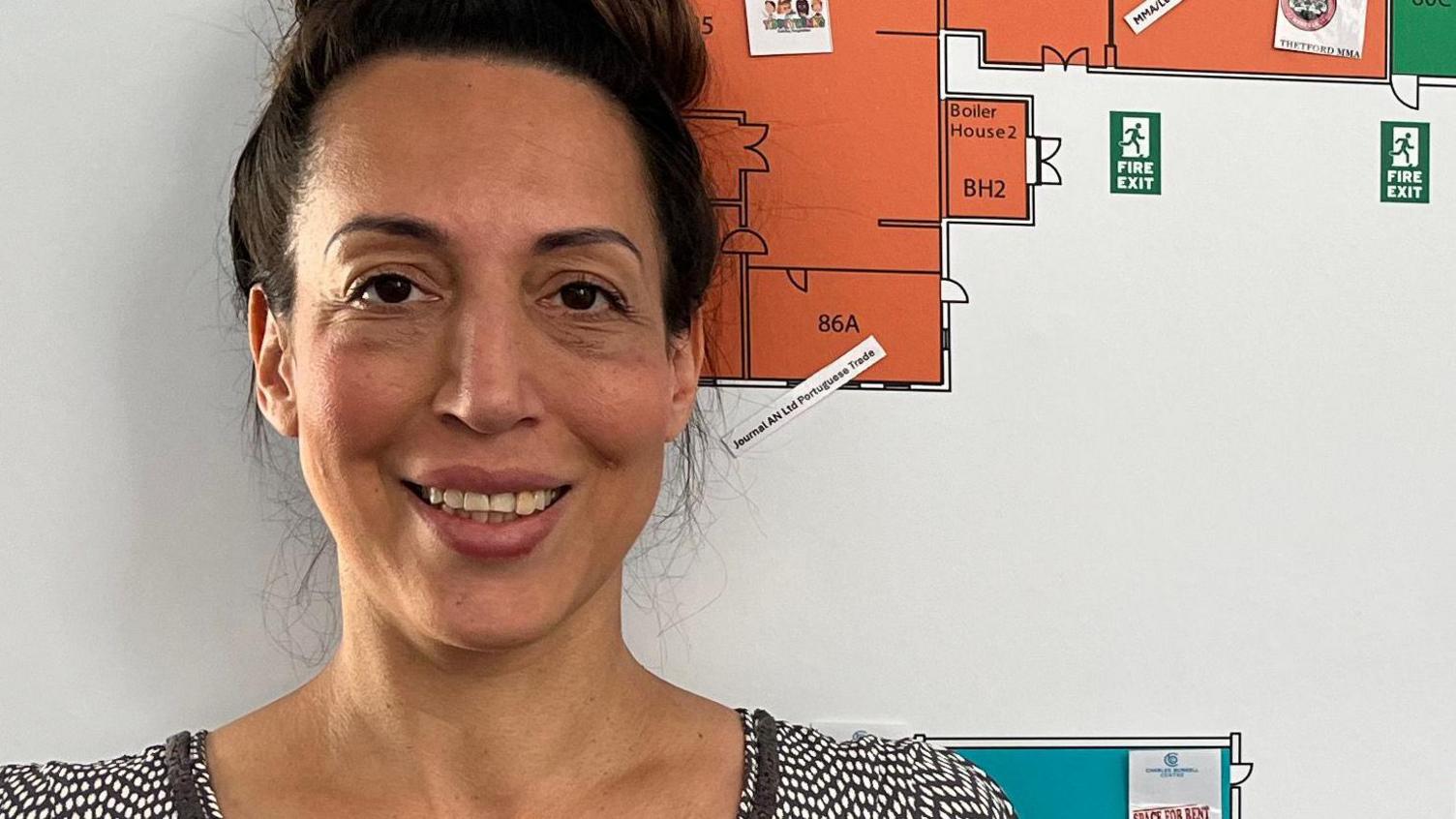 Nicola Welham is the centre manager of The Charles Burrell Centre a former high schools turned into community and business hub. She is wearing a black and white dress and has her brown hair in a bun. She is standing next to a map of the centre