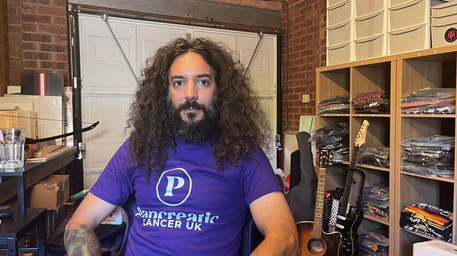 A man in a purple T-shirt with the words Pancreatic Cancer UK on the front. He has long dark hair and a beard. There are shelves to the right of him on the photo.