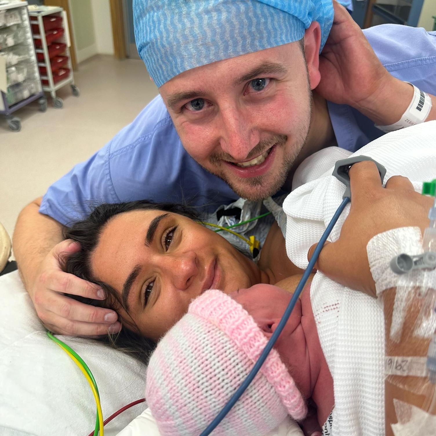 Aisha McCracken with baby after giving birth. Her husband is leaning over her and is wearing blue theatre scrubs. The baby is on top of her chest and has a pink and white woolly hat on. The couple are smiling at the camera and there are tubes coming out of Aisha's arm.