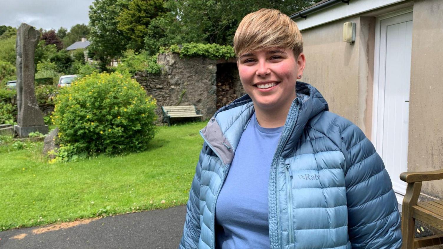 Cara Evans stood smiling at the camera, she is wearing a blue coat and t-shirt. Behind her is a green bush and a grassy verge.