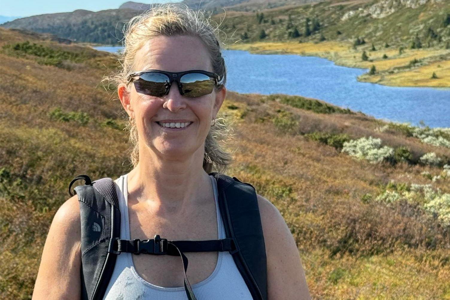 Frøydis Adamson standing outside on heath land, by a lake - wearing sunglasses, a short sleeve top and rucksack straps are visible. She has blond hair and is smiling.