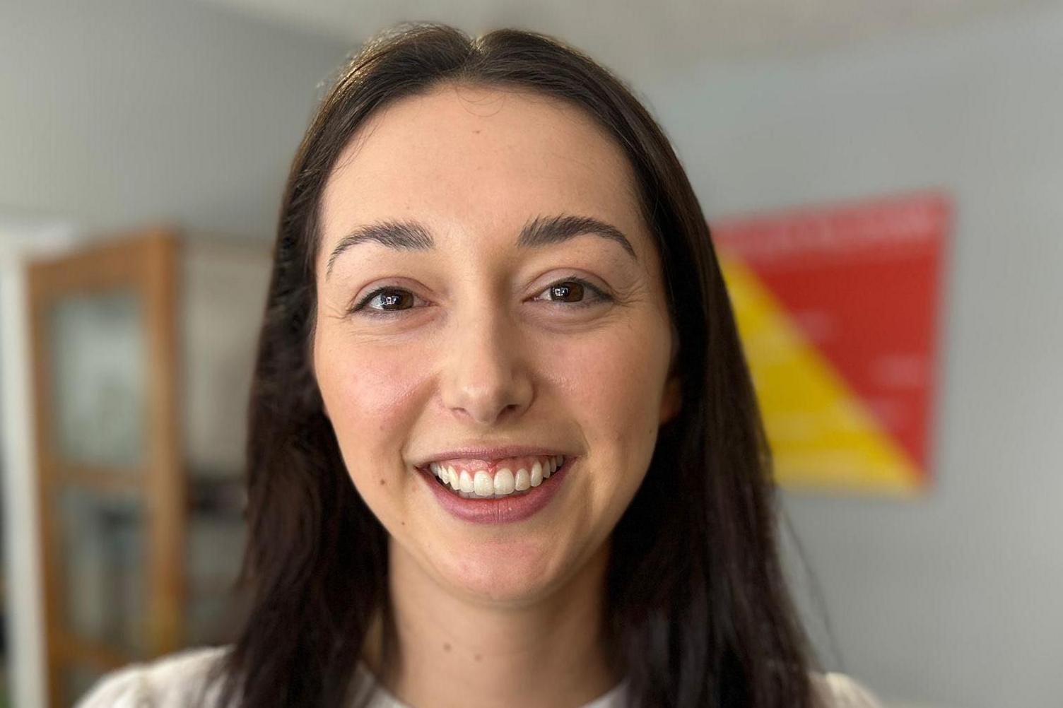 A woman with dark hair and brown eyes wearing a white top smiles into the camera