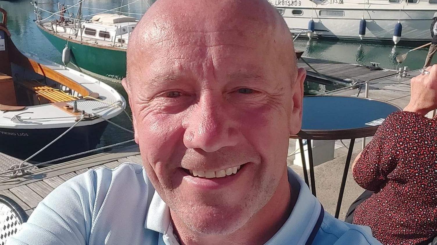 A bald man smiles at the camera. He is sitting next to a marina with small boats behind him.