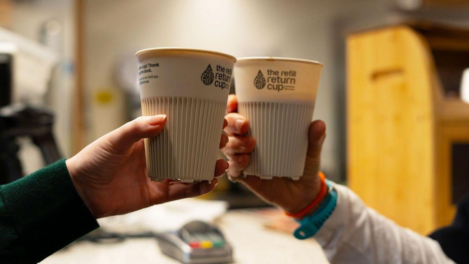 Two people holding white reusable coffee cups. The cups are printed with a graphic reading "The Refill Return Cup". They are holding the cups up together.