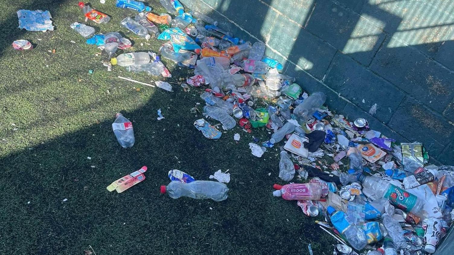Water bottles, such a pink Boost bottle and a Oasis juice bottle, strewn across astro turf. There are also items such as packaging for a vape and a Red Bull can.