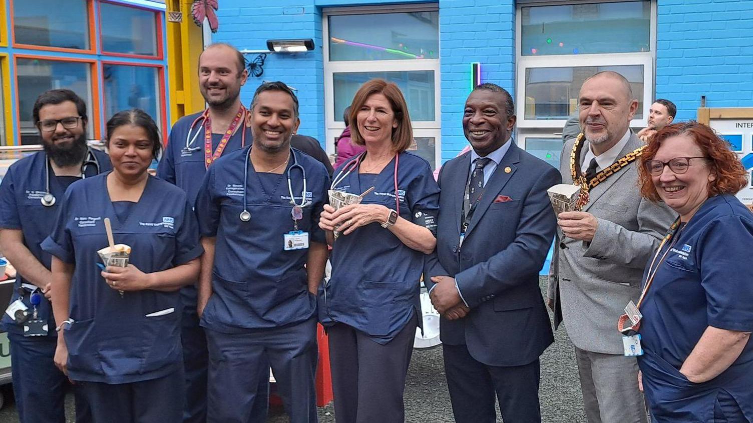Members of hospital staff wear dark blue uniforms, with the Mayor of Wolverhampton second left in a grey suit and wearing his chains of office. The are standing in the revamped area which is painted mainly blue