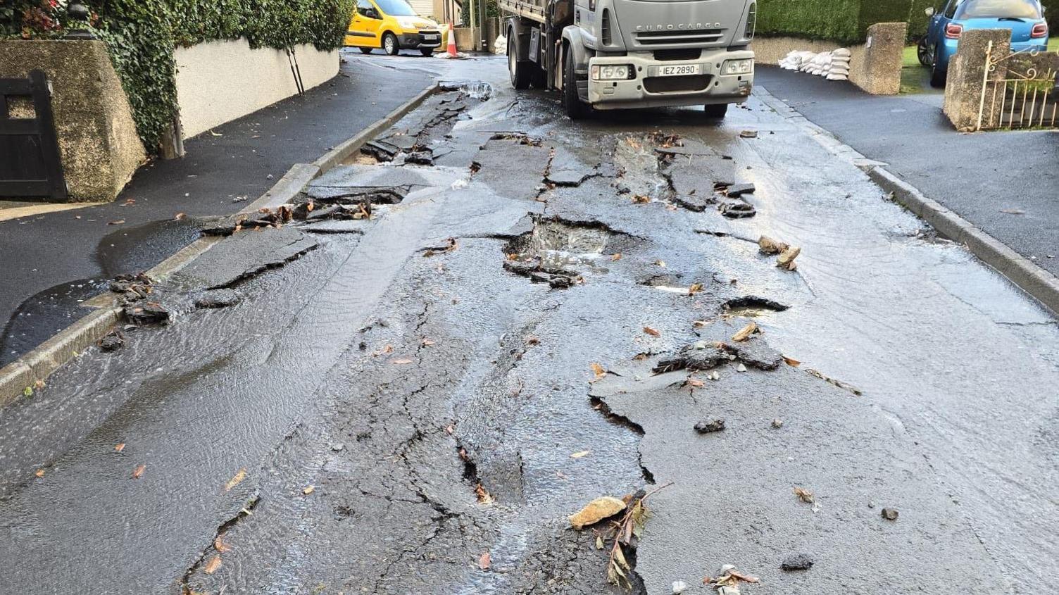 A section of black tarmac has been torn up. There are houses either side of the streets. Small bits of debris are scattered on the road. A grey truck sits at the top of the road.