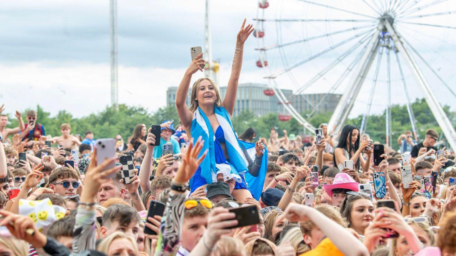 A crowd of people with their hands in the air and people hoisted onto shoulders. One girl on someone's shoulders in the foreground wears all blue with a saltire flag draped around her neck. A Ferris wheel can be seen in the background.