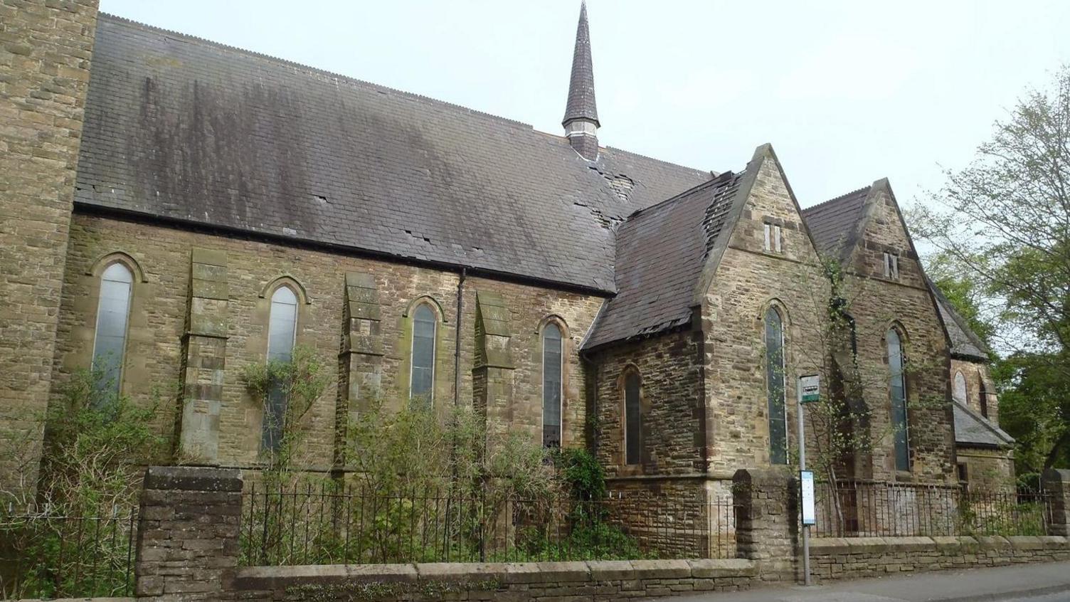 St Aidan’s Church is a tall stone building. Tiles are visibly missing from the roof. The windows that line the building are tall and arch-shaped. Green trees are overgrown out front.