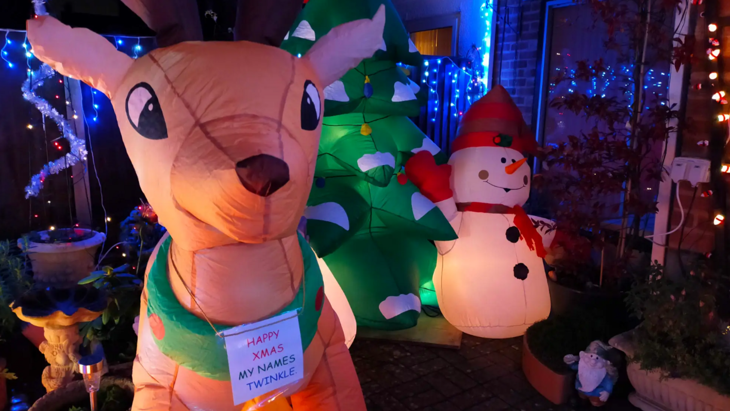 An inflatable reindeer with a sign around its neck reading "Happy Xmas my name's Twinkle" stands next to an inflatable Christmas tree and an inflatable snowman.