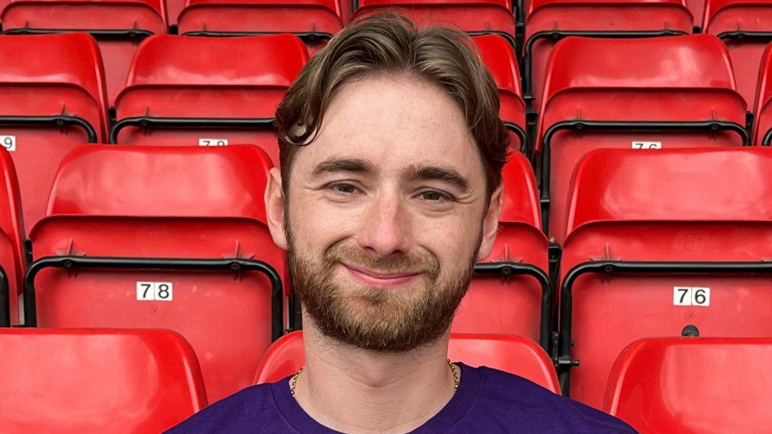 A man with dark hair and a beard is looking at the camera. She is at Walsall's Bescot stadium and red seats are behind her.