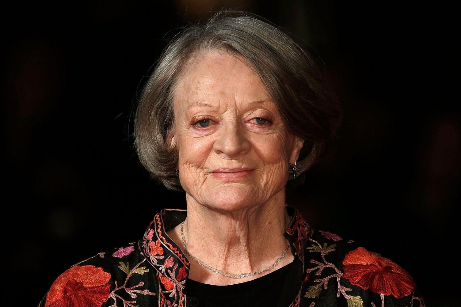 Dame Maggie Smith photographed in 2015, aged 80, against a black background. She is smiling and wearing a black top, a simple necklace and a collar-less floral jacket.