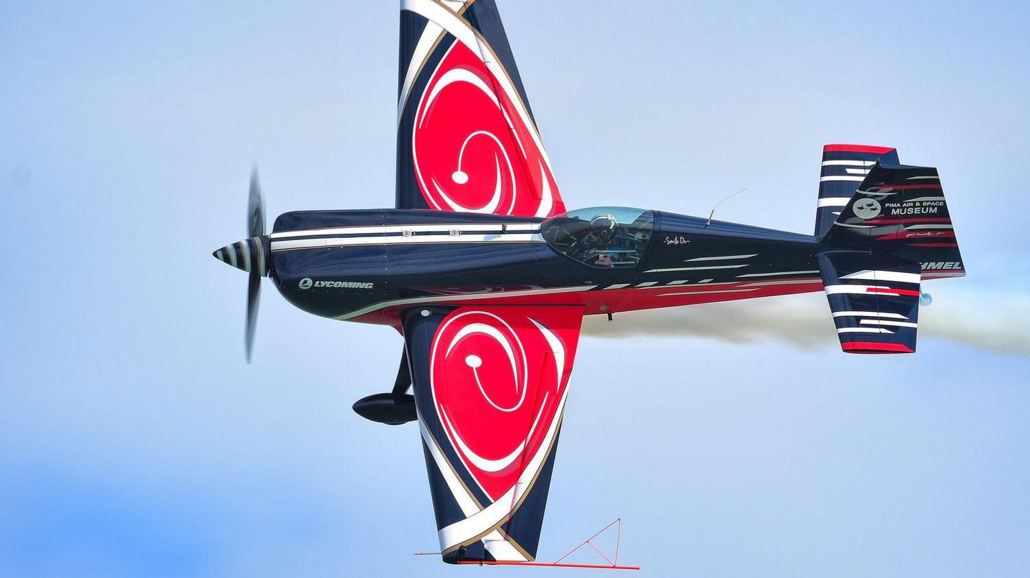 A blue and red one-seater aeroplane flying with fumes trailing off the back