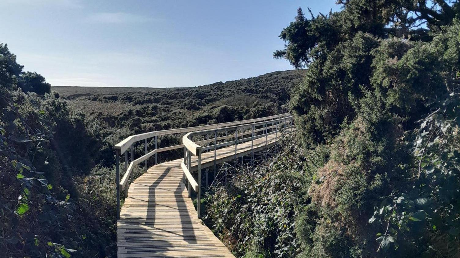 A modern wooden path which winds around it is sits above a hilly terrain, on a clear blue day.
