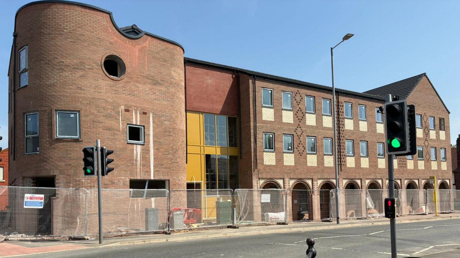 A three-storey brown brick building with a curved corner. There are arches at street level and two storeys of rectangular windows. The building is still under construction and it is behind metal fencing.