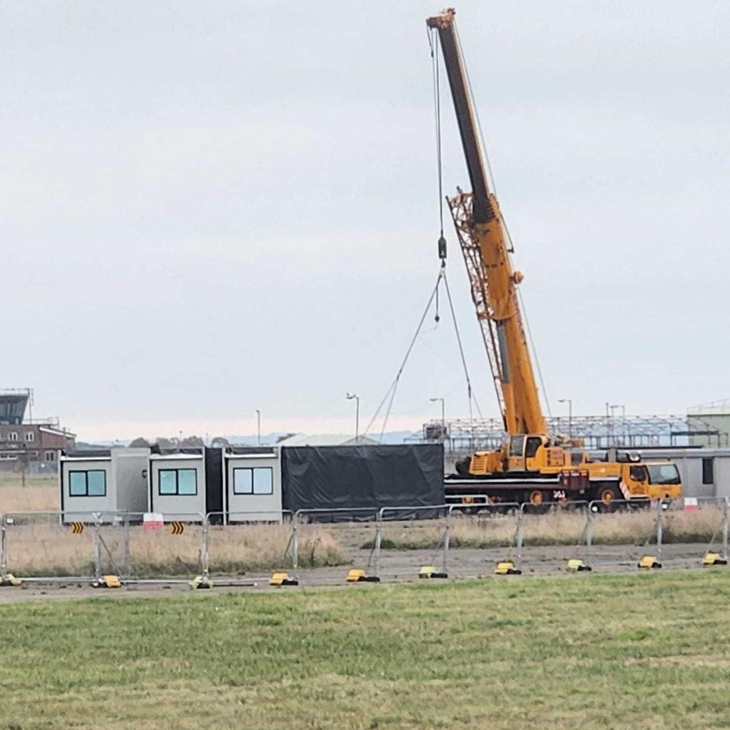 A distance shot of a yellow crane attached to a cabin. There are three cabins in total. They are surrounded by metal fencing and there is grass in front, closer to the screen. 