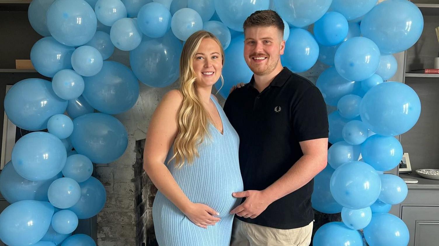 Pregnant blond woman in blue dress standing next to a brown haired man in a black top in front of some balloons