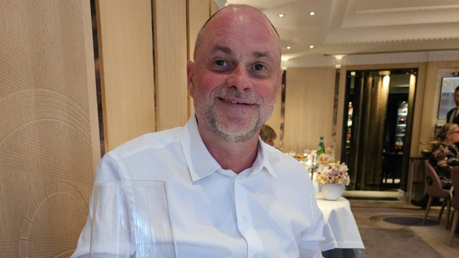 Mark Redmond out at a restaurant. He is a balding man with a beard wearing a white shirt. He is smiling and looking at the camera. It looks fancy and dimly lit. We can see there are people behind him. 