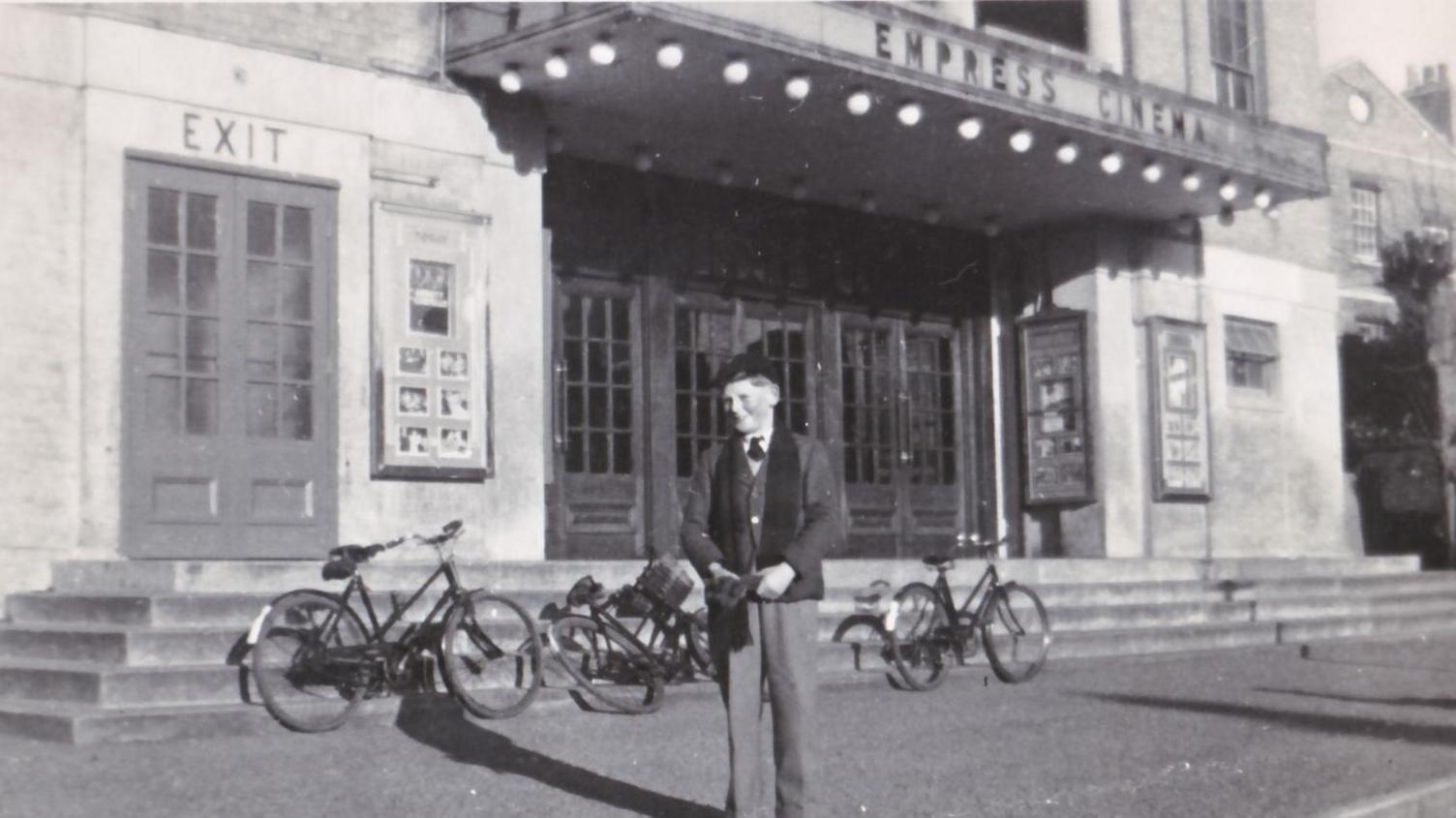 A black and white photo of a smart boy in a suit, with a school cap and scarf around his neck, with three bikes near some steps leading up to the entrance of an old cinema.