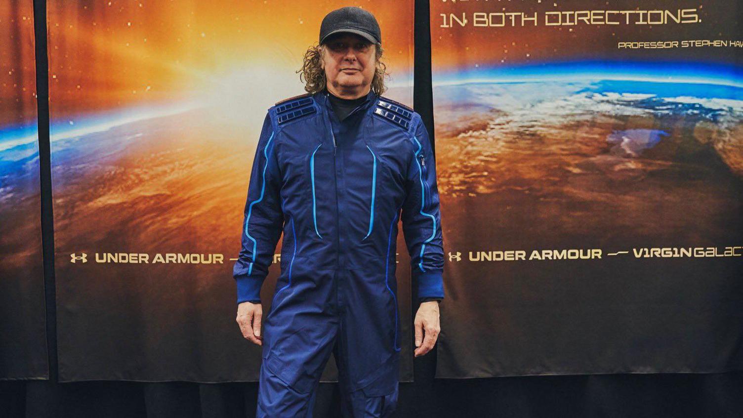 A middle aged man in a blue space suit. He has long hair and is wearing a black cap. He is stood in front of a wall with a picture of planet earth on it.