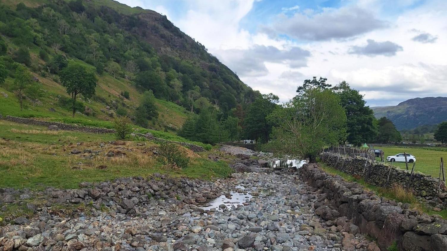 A section of the Upper Derwent. Much of the riverbed is dry with stones visible along a large stretch. There are fields to either side along with hills and trees. The sky is blue but with some areas of cloud.