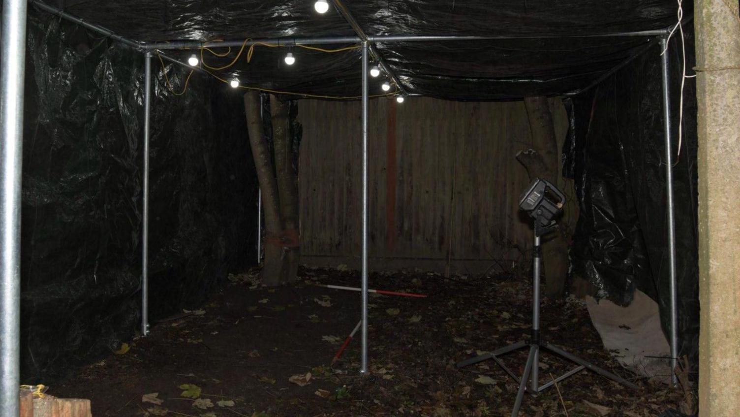 A tent put up over the site in the garden where Abiyah's body was found. There are metal poles holding up the structure, which is covered with a tarpaulin, and there are lightbulbs attached to railings overhead, and there is equipment inside.