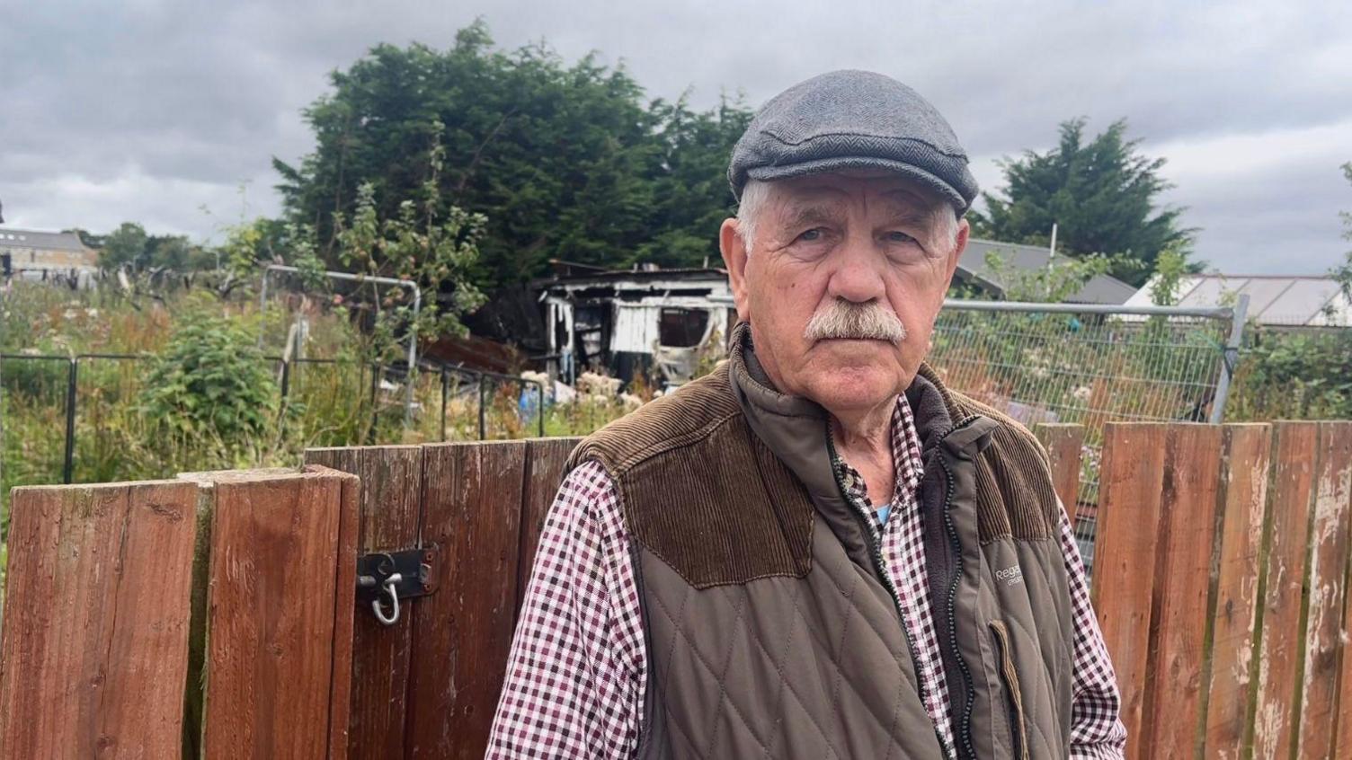 An older man wearing a grey peaked cap, brown bodywarmer and check shirt. He has a grey moustache and stands with his back to a wooden fence behind which are allotments. 