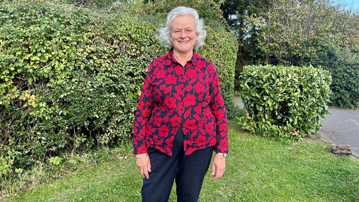 A woman in a red flowery blouse and black trousers. She is standing on a garden and there are hedges behind her.