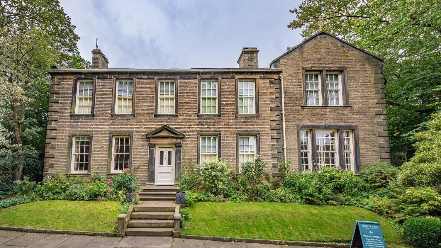 The Brontë Parsonage Museum. A large detached house with white doors and windows. The front gardens are green with wide stone steps.