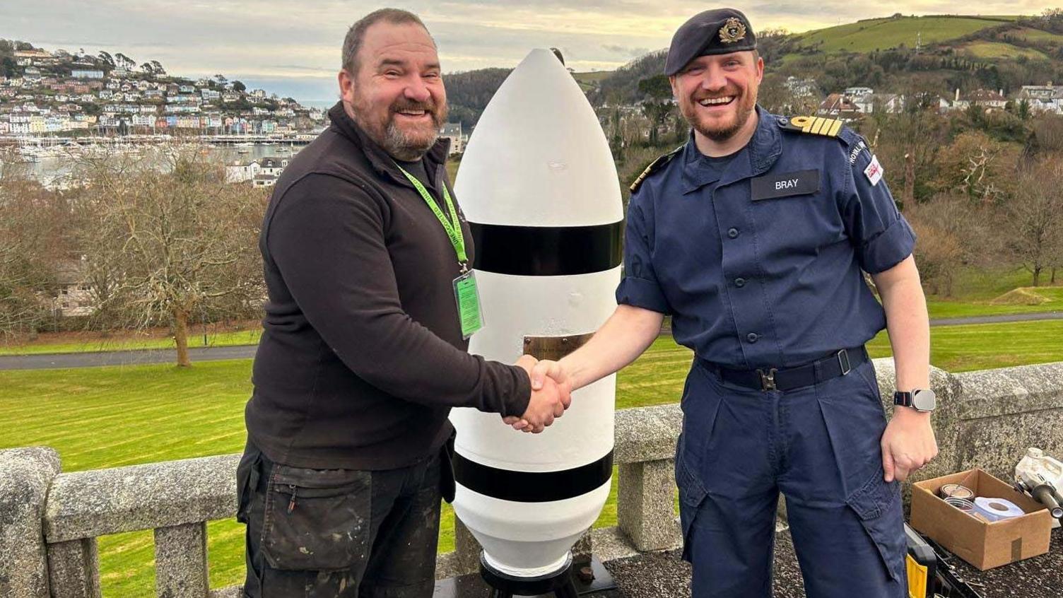 Dressed in black, master wheelwright Greg Rowland shakes the hand of Captain Andy Bray, who is dressed in a uniform of a dark blue short-sleeved shirt and trousers, plus a naval beret with badge on it.
They are standing in front of large white bomb shell, which is half the height of Mr Roland. It has a plaque on its casing. The town of Dartmouth is in the background.