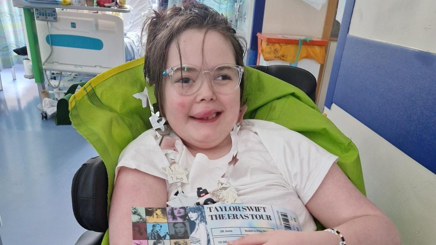 A little girl with brown hair tied back and glasses, smiling holding Taylor Swift tickets