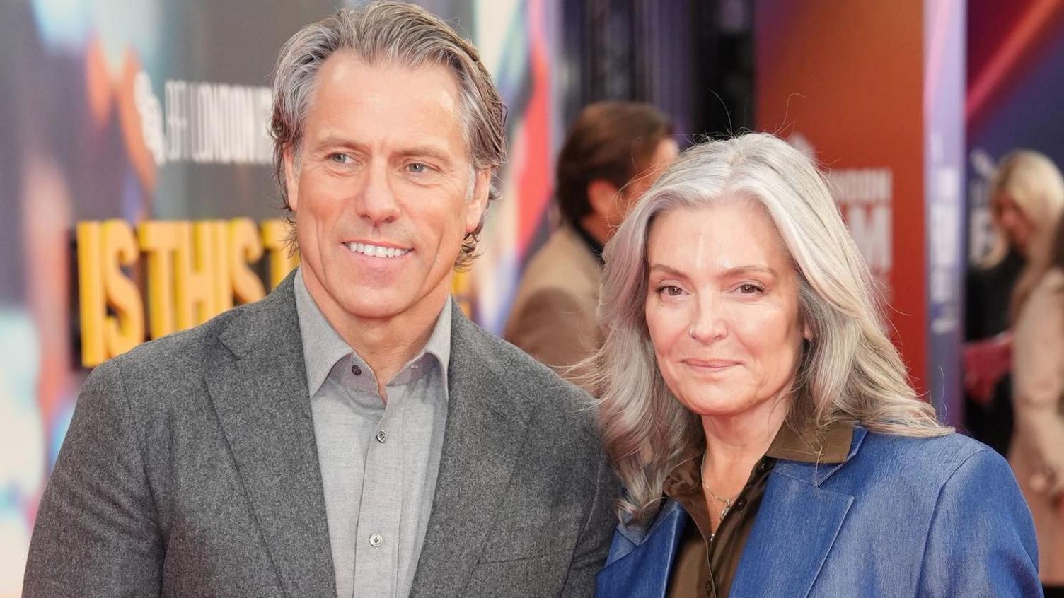 John and Melanie Bishop attending the screening of Is This Thing On? in London. John, with grey hair, is wearing a grey suit and lighter grey shirt. Melanie, with longer grey hair, is wearing a blue jacket and brown shirt. They are both smiling.