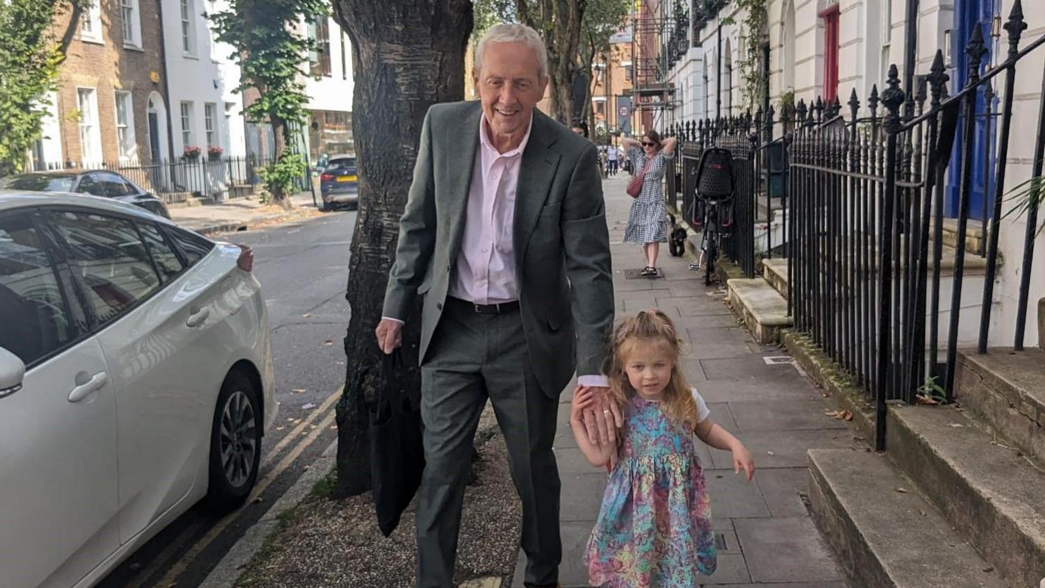 Stan, who wears a grey suit with a light pink shirt, leans down to hold Maggie's hand as they walk down a London street of terraced townhouse. Maggie is wearing a rainbow-coloured dress decorated with butterflies.