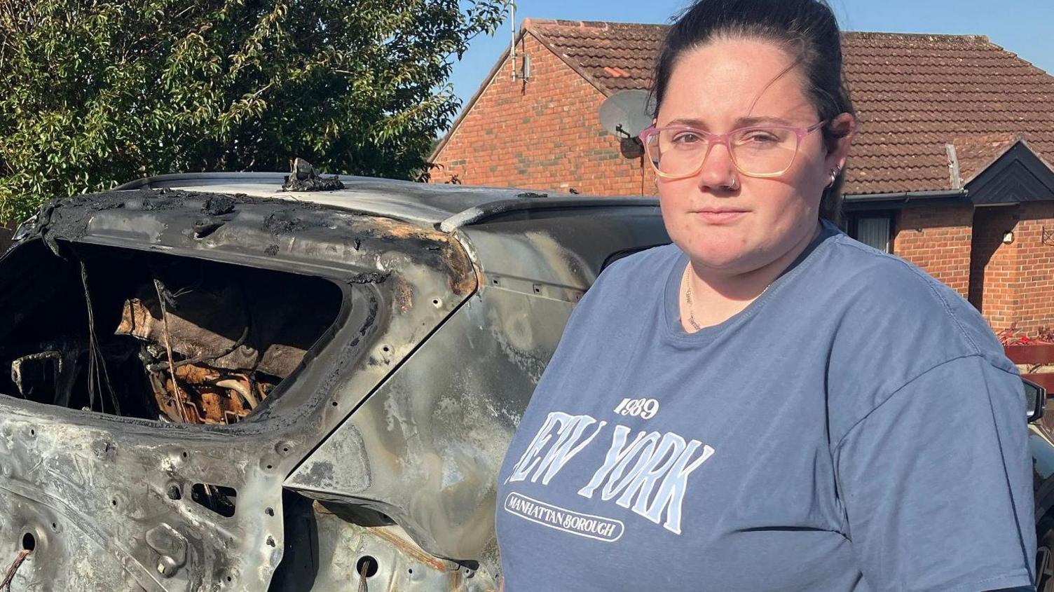 Leonie McLaughlin holds a walking stick as she stands in front of a burned out car. She is wearing glasses and a grey/blue t shirt.