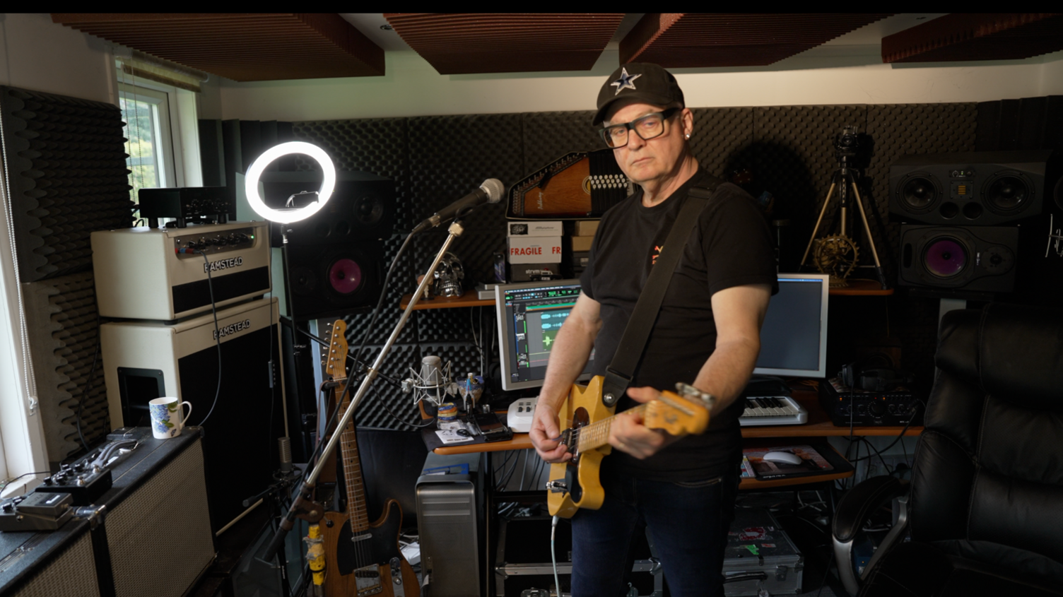 Singer and guitarist Kevin Armstrong pictured with his guitar at his home in Hastings. Kevin is wearing all black, including a black cap. A ring light can be seen in the background alongside amps and recording equipment.