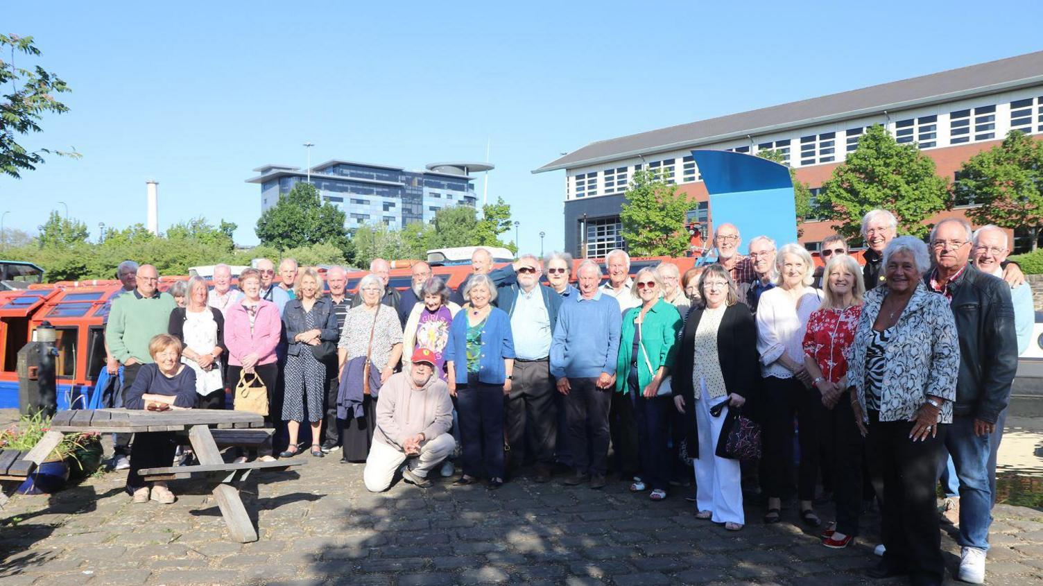 A group of about 40 people who were part of a ten pin bowling club in the 1970s stand next to a canal boat.