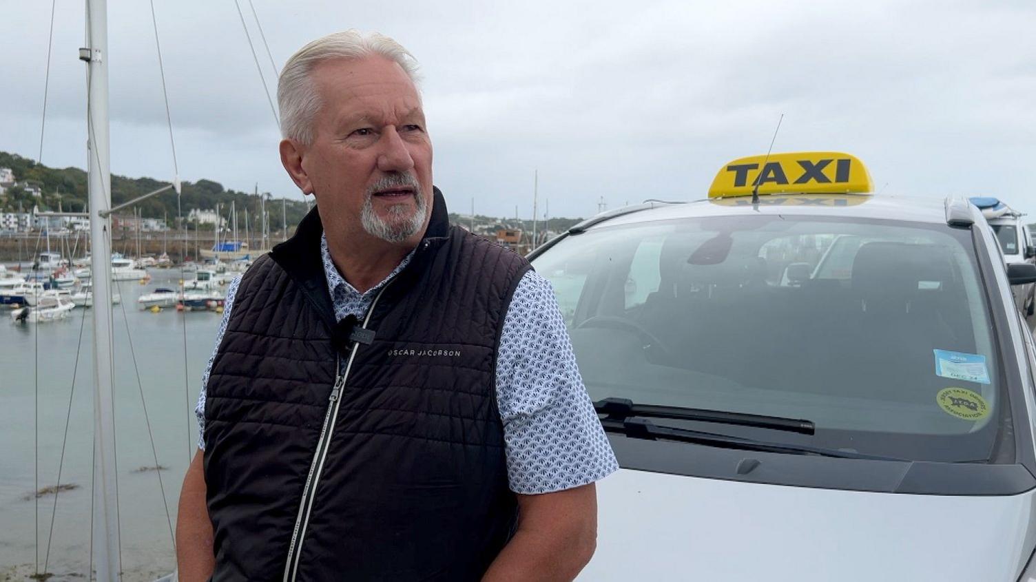 Mick Tostevin, of the Jersey Taxi Drivers Association, standing outdoors next to a white vehicle with a "taxi" sign on top. He has white hair, a moustache, and a grey goatee beard. He is wearing a short-sleeved, light-coloured patterned shirt under a dark puffer vest. Behind him, is a marina with boats 