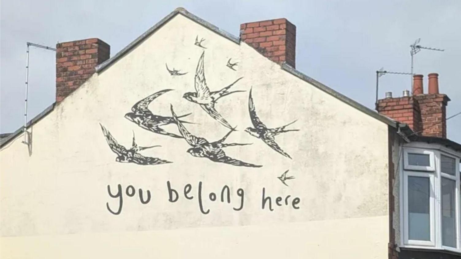 A piece of public artwork on the side of a semi-detached house. It shows a flock of birds and reads: "You belong here" 