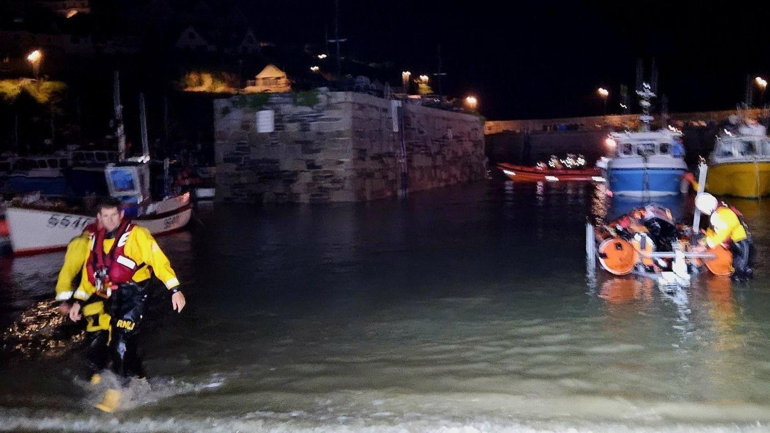 Three members of the Newquay RNLI crew wade back to shore in the town's harbour following a rescue. All three crew are wearing black and yellow overalls and red lifejackets. One of the crew is making an adjustment to an orange rib boat.