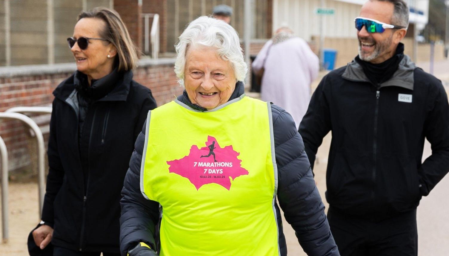 Chris Bryer-Ash's grandmother walking on Bournemouth seafront wearing a luminous yellow vest with the seven marathons seven days logo 