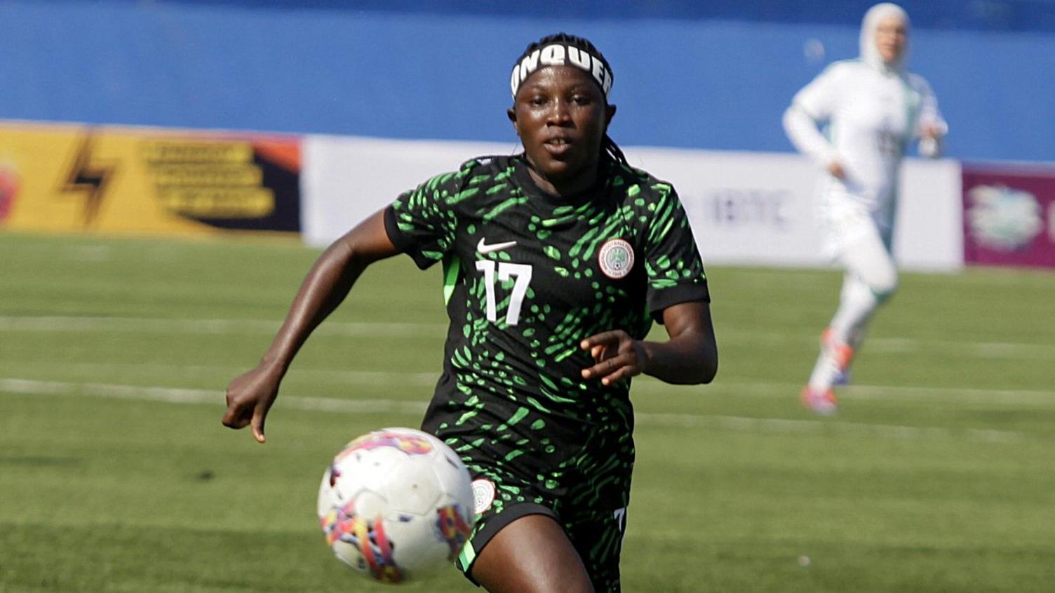 Blessing Illivieda in green and black Nigeria kit and a wearing a distinctive headband with writing on it runs towards the camera while chasing a football that is bouncing in front of her during a match