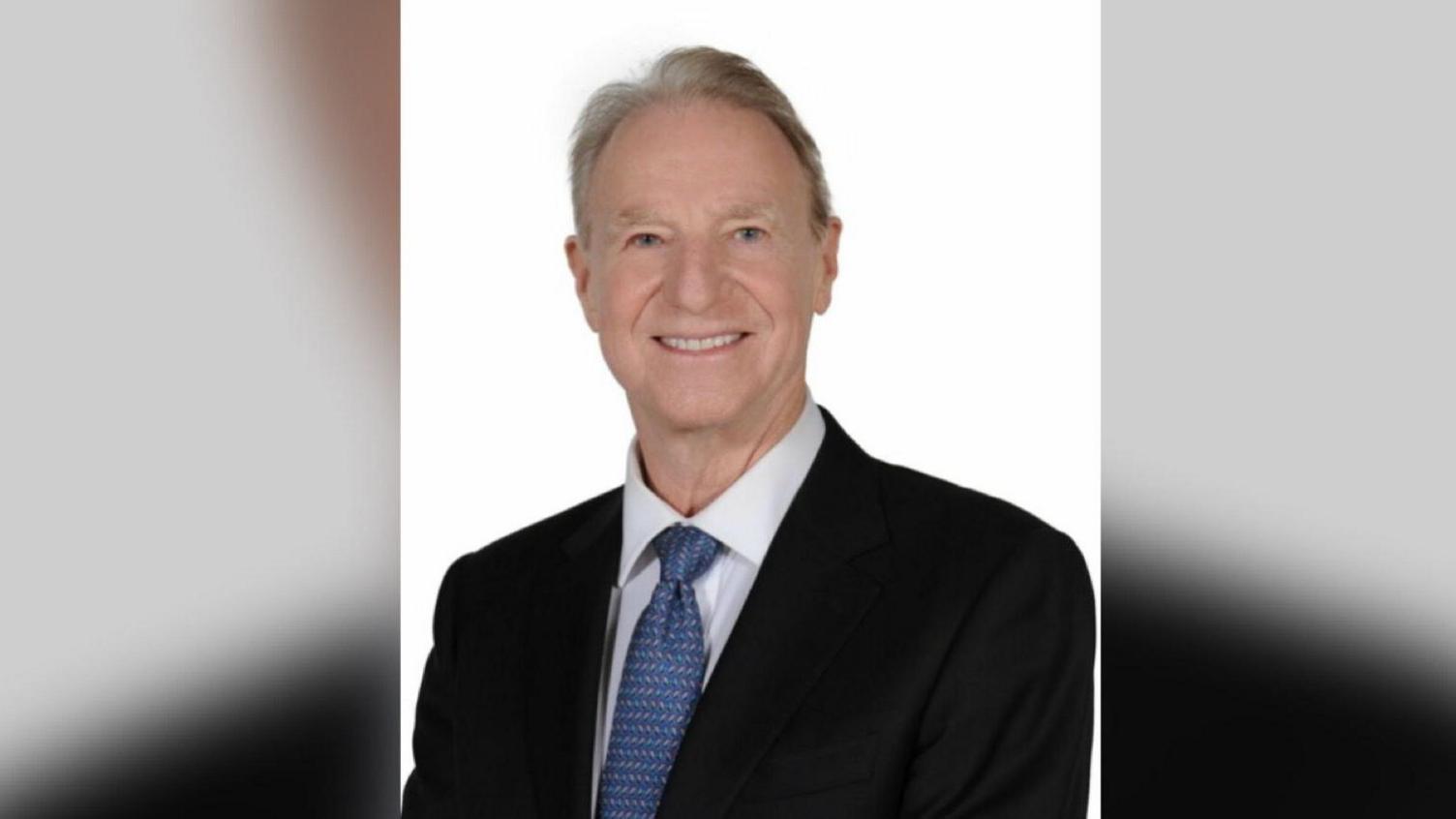 Ian Smith looks into the camera. He is wearing a suit with a blue tie and has grey and brown hair.