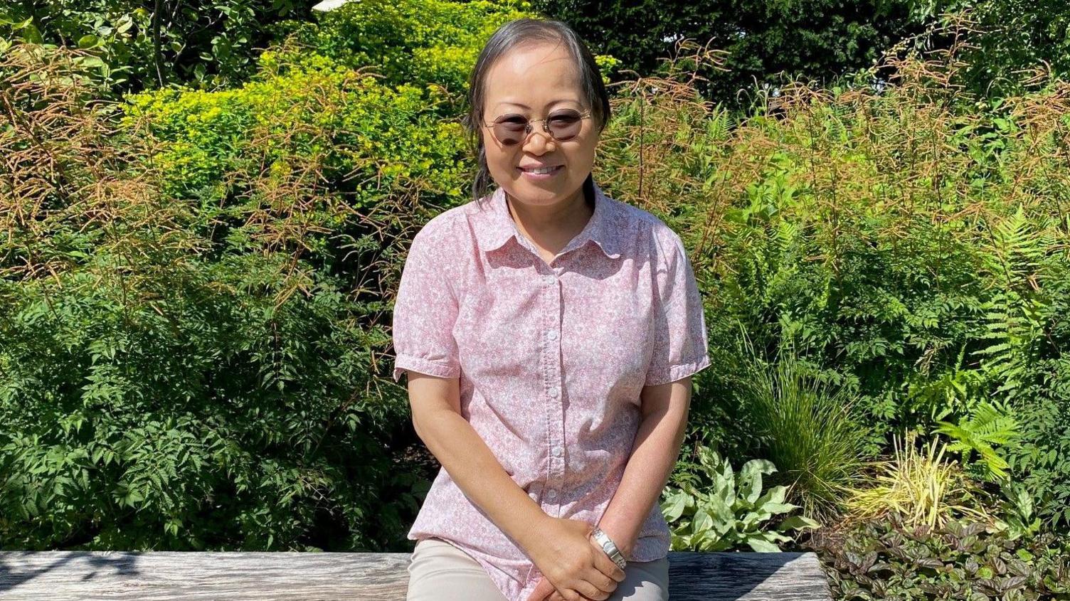 Sophie, sat in a sunny garden at Maggie's in Manchester. She is wearing sunglasses, light-coloured trousers and a short-sleeved pink shirt. She is sat on a wooden bench and there are plants and shrubs behind her.