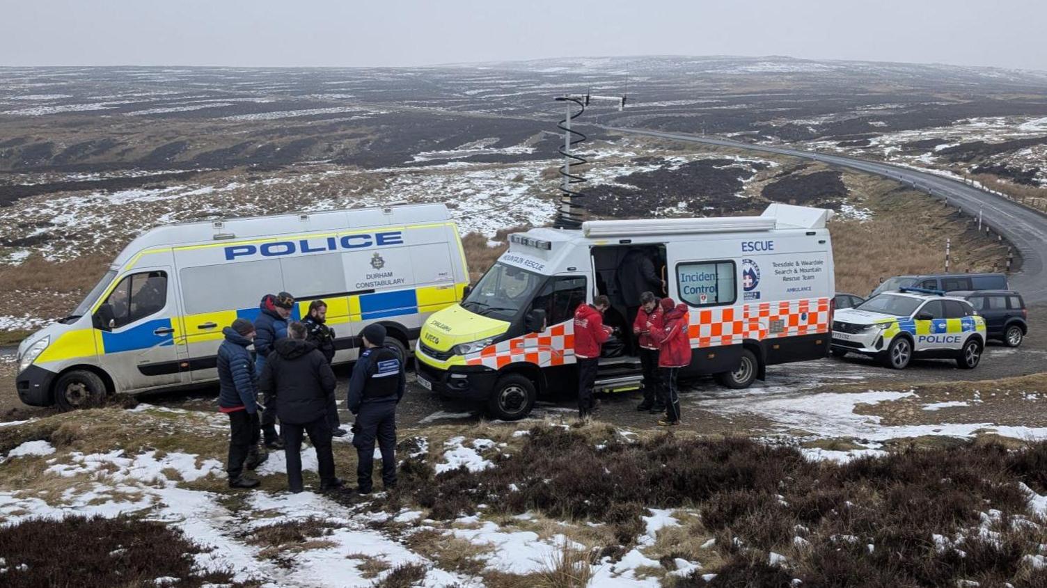 A police van parked alongside a mountain rescue van. Mountain rescuers can be seen in front of the vans in red jackets with police officers in black standing in a separate group. Moorland can be seen behind the vans, which stretches beyond the horizon.