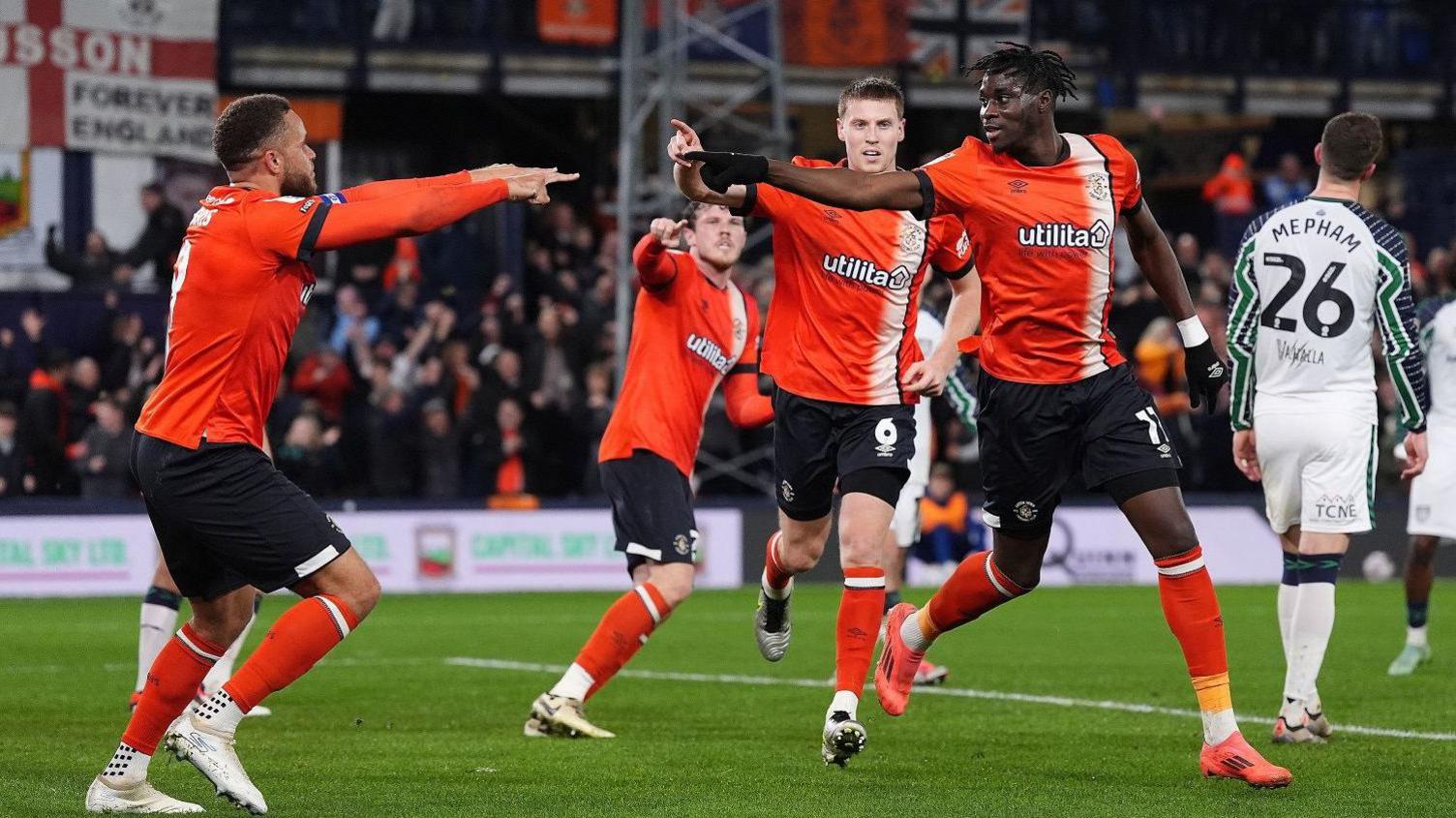 A group of four Luton Town footballers celebrate a goal win on the pitch at Kenilworth Road by high fiving each other. they are wearing their home kit, orange "utilita" sponsored T-shirts and socks with black shorts. The crowd is blurred in the background and a Sunderland player is seen from behind wearing a white kit and the number 26.