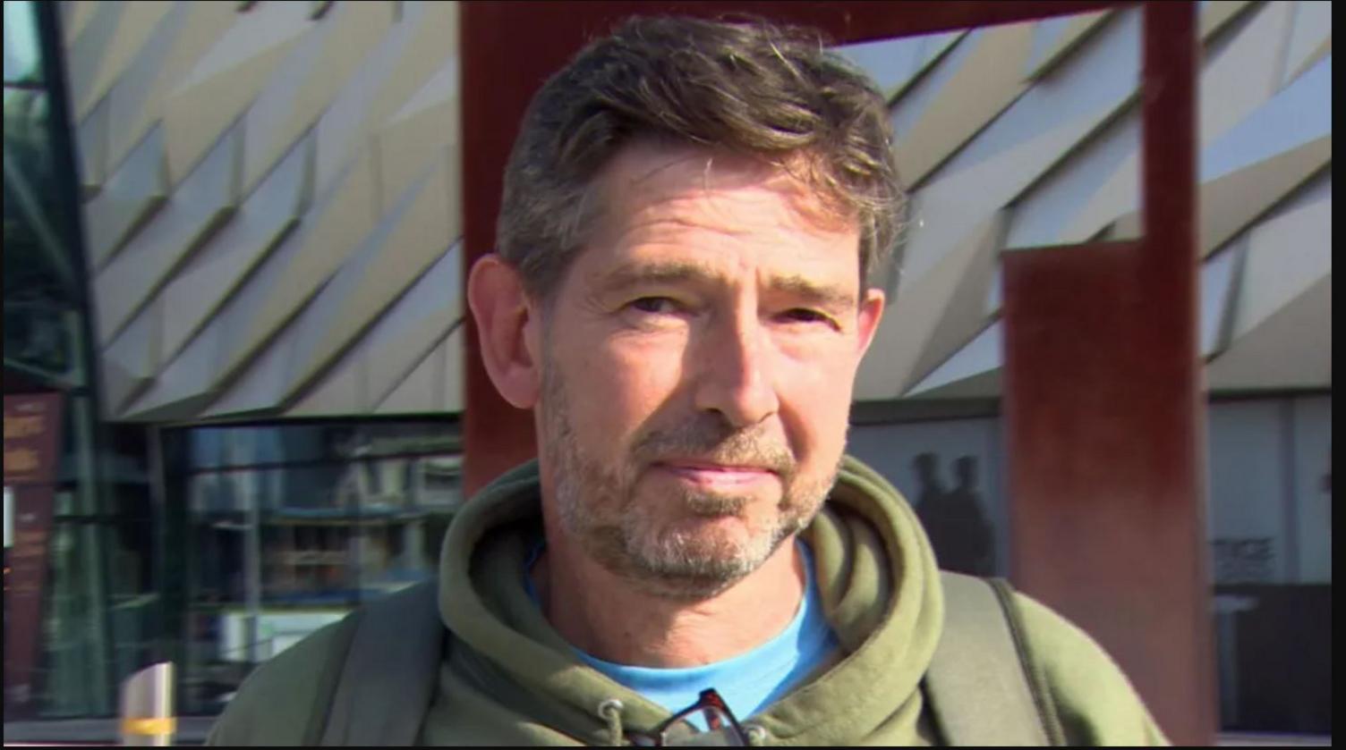 Man with brown hair and beard wearing a green hooded jacket in front of the Titanic Museum in Belfast 