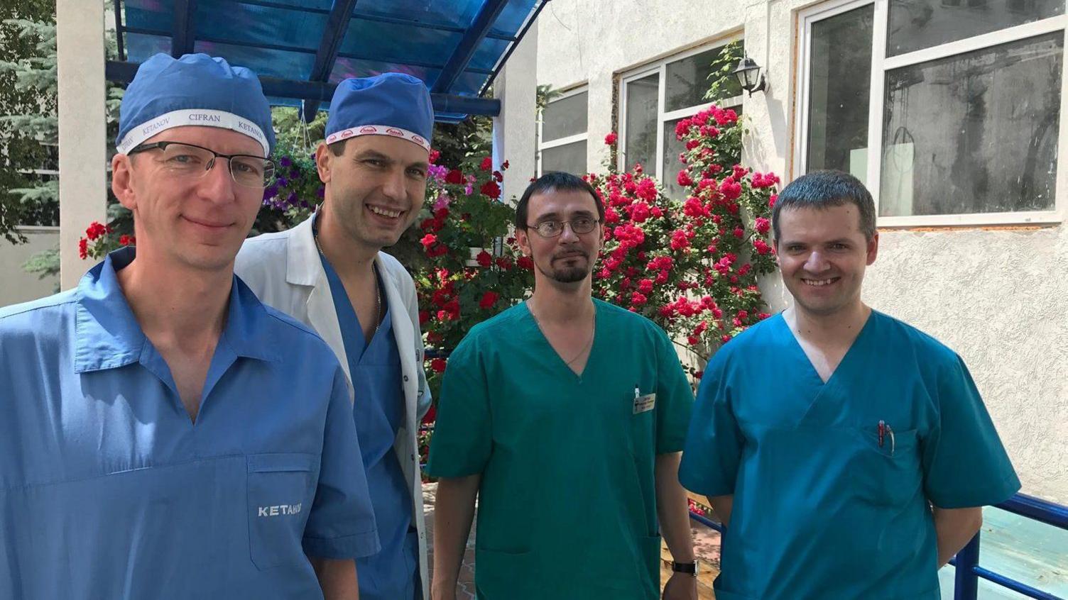 Four men stands outside a hospital wearing scrubs. Leo is on the left, wearing light blue scrubs and a matching cap and thin wire framed glasses. To the right, a man with black hair wears pale blue scrubs, a blue cap and a white overcoat, a man stands next to him with black short hair and a black beard, wearing deep green scrubs and another man stands next to him, with matching blue scrubs on. They are all smiling at the camera