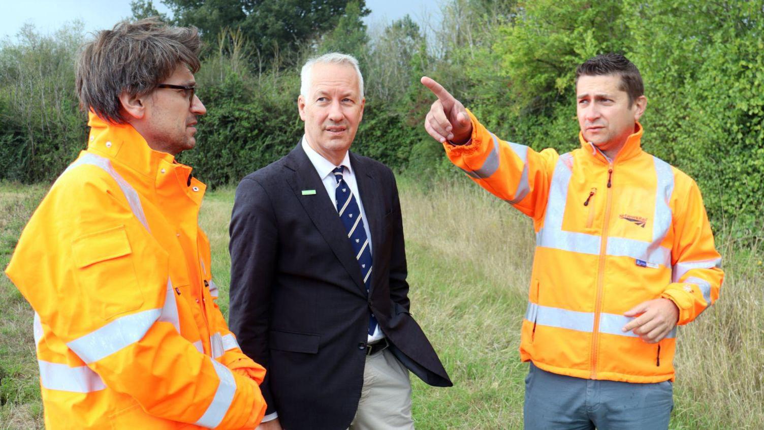 Two men in orange high visibility jackets stand either side of Gideon Amos MP in a navy blazer and tie. They are in a field and one of the men in orange is pointing towards something out of shot