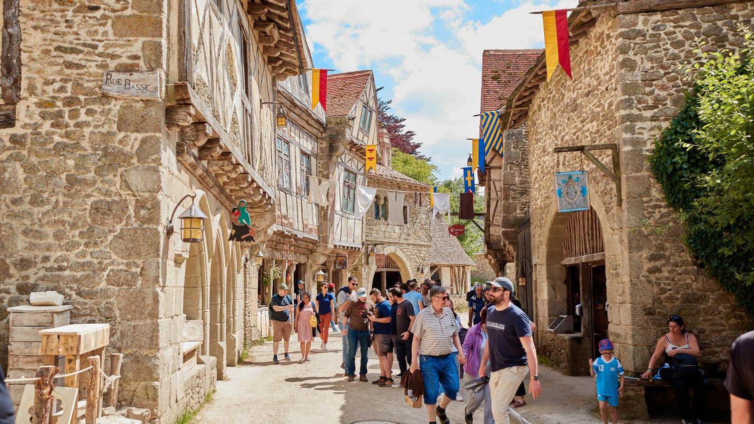 A medieval village scene complete with stone and Tudor-looking buildings with exposed wooden beams. There are tens of modern people walking down the street.